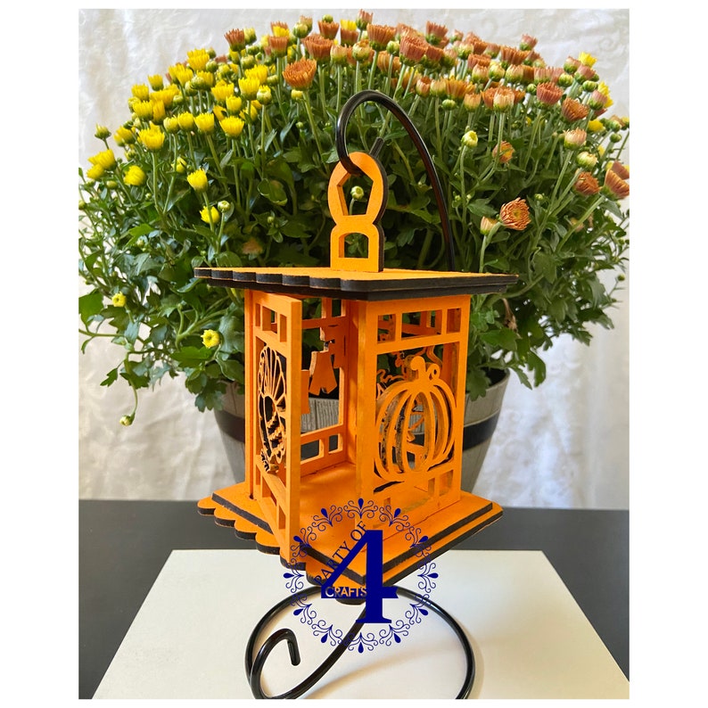 May include: Orange wooden lantern with a black metal stand. The lantern has cutouts of a pumpkin, spider web, and a ghost. The lantern is sitting on a stand with a black metal base and a black metal scroll design.