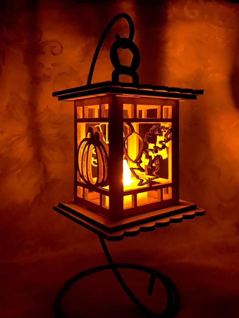 May include: A wooden lantern with a black metal stand. The lantern has a carved pumpkin and other Halloween-themed designs. A candle is lit inside the lantern, casting a warm glow.