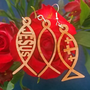 May include: Three wooden fish-shaped earrings with a religious theme. The earrings are made of light brown wood and have a delicate design. One earring features the word "JESUS" carved into the fish, another has a cross carved into the fish, and the third has a simple fish design.