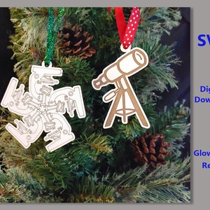 Puede incluir: Dos adornos navideños, uno con forma de copo de nieve y el otro con forma de telescopio, cuelgan de un árbol de Navidad verde. Los adornos son blancos y tienen un diseño detallado.