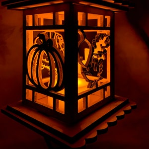 May include: A wooden lantern with a light inside, featuring a pumpkin and a turkey cut-out design. The lantern is lit and glows a warm orange color.