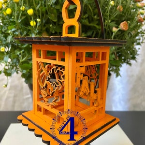 May include: Orange wooden lantern with a black trim and a cutout design of pumpkins and the word "FALL". The lantern is hanging from a black metal stand. The bottom of the lantern has a scalloped edge and a blue circle with the text "Party of 4 Crafts".