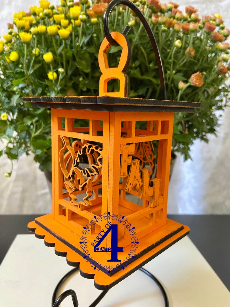 May include: Orange wooden lantern with a black trim and a cutout design of pumpkins and the word "FALL". The lantern is hanging from a black metal stand. The bottom of the lantern has a scalloped edge and a blue circle with the text "Party of 4 Crafts".