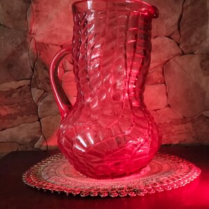 Pode incluir: Uma jarra de vidro vermelho vibrante com um padrão geométrico texturizado e repetitivo em toda a sua superfície. A jarra possui uma alça curva na lateral e uma abertura larga no topo com um pequeno bico, iluminada por uma luz vermelha.
