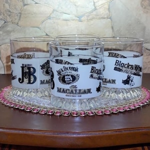 May include: A set of five clear drinking glasses with white labels featuring various brand names like "JB", "Jack Daniel's", "Black & White", and "Macallan". The glasses are arranged on a decorative, oval-shaped coaster with a pink and gold trim.