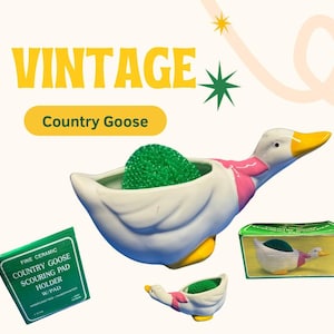 Może przedstawiać: Vintage ceramiczny uchwyt na gąbkę Country Goose z zieloną gąbką. Gęś jest biała z żółtym dziobem i stopami oraz różowym szalikiem. Obraz zawiera oryginalne pudełko i mniejszą wersję uchwytu.