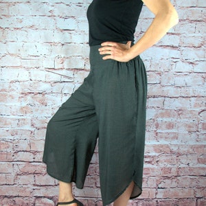 Puede incluir: Una persona con una camiseta negra y pantalones grises oscuros de pierna ancha con un sutil estampado de puntos. Los pantalones tienen cintura alta y una abertura en el dobladillo. Sandalias negras completan el atuendo. El fondo es una pared de ladrillos.