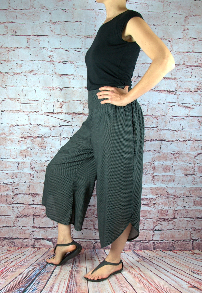 Puede incluir: Una persona con una camiseta negra y pantalones grises oscuros de pierna ancha con un sutil estampado de puntos. Los pantalones tienen cintura alta y una abertura en el dobladillo. Sandalias negras completan el atuendo. El fondo es una pared de ladrillos.