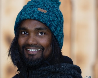 Può includere: Un uomo con lunghi capelli neri indossa un berretto di lana blu con un pon pon in cima. Il berretto ha un'etichetta in pelle marrone con la scritta "Heine" su di essa. Sta sorridendo e guardando la telecamera.