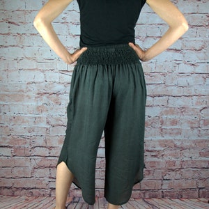 Puede incluir: Pantalones capri grises oscuros con cintura fruncida y dobladillo con abertura. Los pantalones se combinan con una camiseta negra y sandalias. El fondo es una pared de ladrillos.