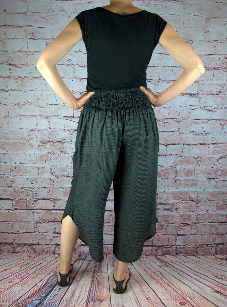 Puede incluir: Pantalones capri grises oscuros con cintura fruncida y dobladillo con abertura. Los pantalones se combinan con una camiseta negra y sandalias. El fondo es una pared de ladrillos.