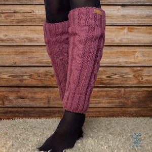 Op de afbeelding: Paar mauve gebreide beenwarmers met een kabelpatroon. De beenwarmers hebben een geribbelde manchet en een klein leren label. De achtergrond is een houten muur. Ontworpen om op de onderbenen te dragen.
