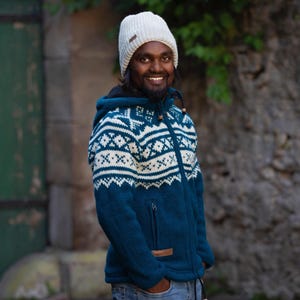 Può includere: Un uomo indossa un maglione blu e bianco con un motivo a maglia con un cappello di lana bianco. Il maglione ha una cerniera e un cappuccio.