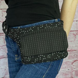 May include: Black fanny pack with a geometric pattern and white star accents. The pack has multiple pockets and a buckle closure. The fanny pack is worn around the waist over blue jeans.