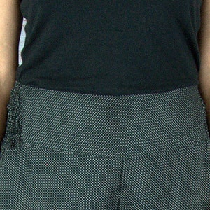 Puede incluir: Una camiseta sin mangas negra combinada con pantalones de cintura alta de color gris oscuro con un peque&ntilde;o estampado de lunares blancos. Los pantalones tienen una cintura ancha y un detalle de flecos en el lateral. El fondo es una pared de ladrillo.