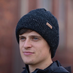 Könnte beinhalten: Eine dunkelgraue Strickmütze mit einem kleinen braunen Etikett. Die Mütze wird von einer Person mit blauen Augen getragen.
