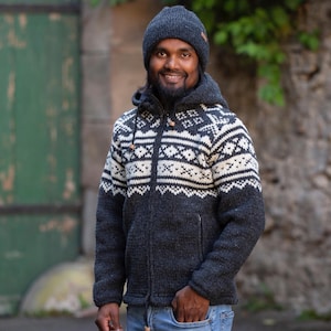Può includere: Una giacca di maglieria grigia scura con un motivo geometrico bianco e nero. La giacca ha una cerniera e un cappuccio. La persona indossa un berretto grigio scuro.