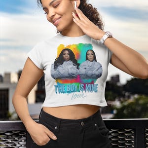 May include: White crop top featuring a colorful graphic of two women and the text "THE BOY IS MINE Tour". The top is worn with black pants. The image is taken outdoors.