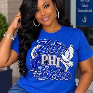 Könnte beinhalten: Ein königsblaues Kurzarm-T-Shirt mit einem Grafikdruck, der Zeta PHI Beta in metallisch blauer und silberner Schrift zeigt. Das Design umfasst eine weiße Taube, eine Kette aus weißen Perlen und dunkelblaue Rosen mit funkelnden Akzenten.