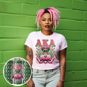 May include: A pink t-shirt with the letters "AKA" in green and pink glitter. The shirt features a pink purse with green jewels and a crown on top. The purse is surrounded by green jewels and pink high heels.