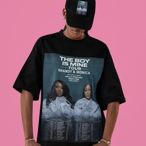 Pode incluir: Camiseta preta com o texto "THE BOY IS MINE TOUR BRANDY & MONICA" e datas da turnê. A camiseta apresenta imagens dos artistas. Um boné de beisebol preto com uma imagem correspondente também é visível.