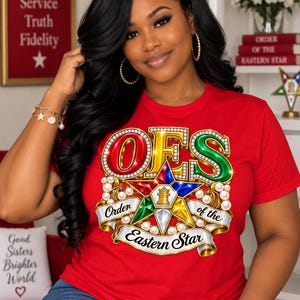 Puede incluir: Una camiseta roja de manga corta con un estampado gráfico del emblema de la Orden de la Estrella de Oriente. El diseño incluye las letras OES en rojo, verde y dorado, una estrella multicolor con una torre central y perlas blancas en una cinta.