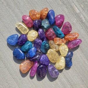 May include: A collection of colorful, tumbled agate stones in various shades of blue, purple, yellow, orange, and pink. The stones are arranged in a circular pattern on a light gray surface.