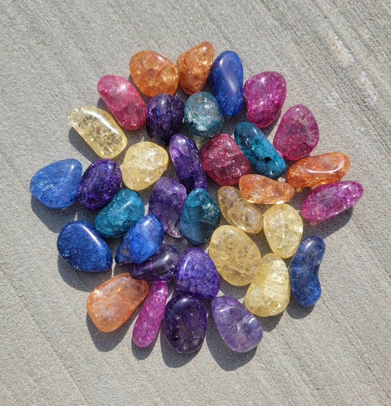 May include: A collection of colorful, tumbled agate stones in various shades of blue, purple, yellow, orange, and pink. The stones are arranged in a circular pattern on a light gray surface.