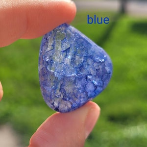 May include: A blue, irregularly shaped, translucent stone with a cracked, crystalline texture. The stone is being held by a finger, and the word "blue" is written in blue letters above the stone.