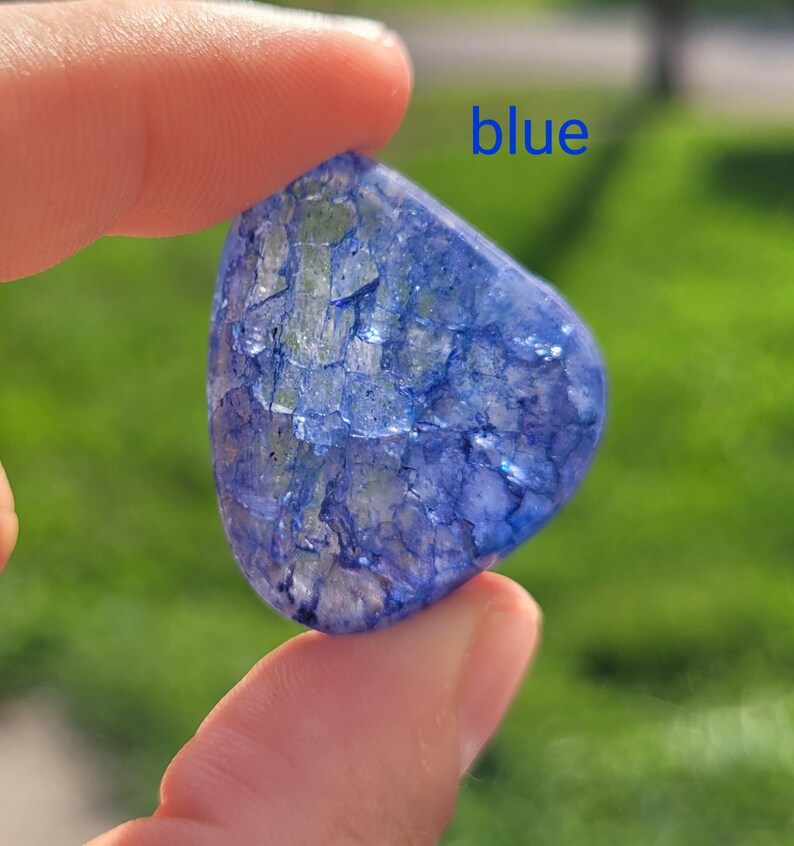 May include: A blue, irregularly shaped, translucent stone with a cracked, crystalline texture. The stone is being held by a finger, and the word "blue" is written in blue letters above the stone.