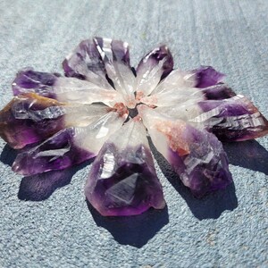 May include: A collection of amethyst crystal points arranged in a circular pattern. The crystals transition from deep purple to clear, with some showing reddish-brown hues. The arrangement is set against a textured gray surface.