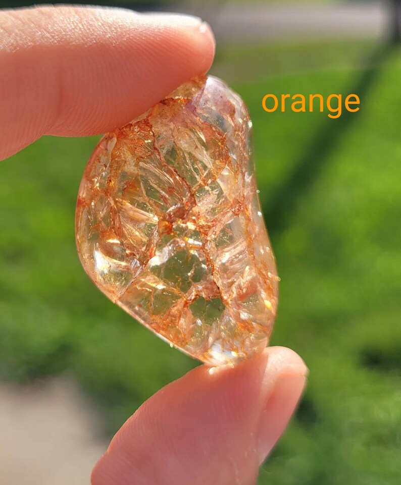 May include: A clear, orange, translucent stone with a unique, intricate pattern of inclusions. The stone is being held in a hand.