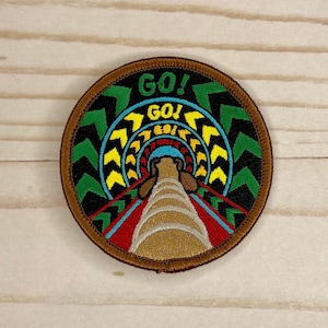 May include: A round embroidered patch with a brown border. The center of the patch features a colorful design of a tunnel with the word "GO!" repeated in a circle. The tunnel is lined with yellow and green chevron patterns.