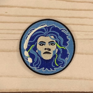 May include: Embroidered patch featuring a blue and white portrait of a person with long blue hair and a white headband. The person has a serious expression and is surrounded by a blue and white wave-like design.