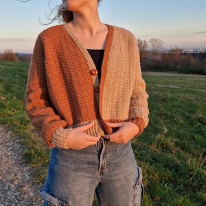 Op de afbeelding: Een tweekleurig gehaakt vest in bruin- en beigetinten, met een V-hals en knoopsluiting. De trui met lange mouwen wordt gecombineerd met een blauwe denim cargobroek. De foto is buiten genomen.