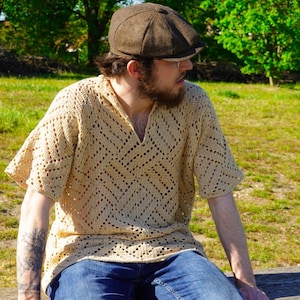 Op de afbeelding: Een beige gehaakt shirt met korte mouwen, een V-hals en een openwerkpatroon. Het shirt wordt gecombineerd met een blauwe spijkerbroek. De persoon op de foto draagt ook een bruine pet.