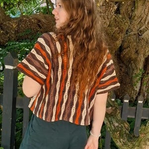 Puede incluir: Una camiseta de crochet de manga corta con un patr&oacute;n de rayas verticales en colores marr&oacute;n, naranja y crema. La camiseta tiene un corte holgado y un cuello redondo. La persona lleva pantalones verde oscuro. El fondo incluye un &aacute;rbol y una valla negra.