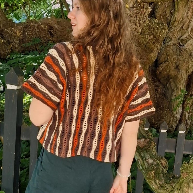 Puede incluir: Una camiseta de crochet de manga corta con un patr&oacute;n de rayas verticales en colores marr&oacute;n, naranja y crema. La camiseta tiene un corte holgado y un cuello redondo. La persona lleva pantalones verde oscuro. El fondo incluye un &aacute;rbol y una valla negra.