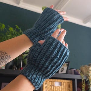 Op de afbeelding: Een paar tealblauwe vingerloze handschoenen, met de hand gehaakt met een geribbeld patroon. De handschoenen worden getoond tegen een achtergrond van een donker tealblauwe muur en een glimp van een plank met decor.