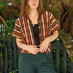 Puede incluir: Un c&aacute;rdigan de crochet de manga corta con botones en tonos marr&oacute;n, naranja y crema. El dise&ntilde;o de tejido abierto revela una camiseta negra debajo. El c&aacute;rdigan tiene cuello en V y se combina con pantalones verde oscuro. El estilo recuerda a los a&ntilde;os 70.