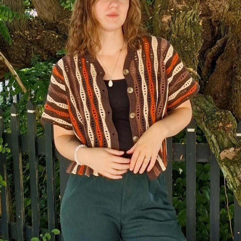 Puede incluir: Un c&aacute;rdigan de crochet de manga corta con botones en tonos marr&oacute;n, naranja y crema. El dise&ntilde;o de tejido abierto revela una camiseta negra debajo. El c&aacute;rdigan tiene cuello en V y se combina con pantalones verde oscuro. El estilo recuerda a los a&ntilde;os 70.