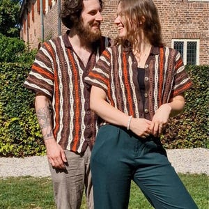 Può includere: Due persone indossano camicie all'uncinetto a righe marroni, arancioni e bianche abbinate, con maniche corte e bottoni. Lo sfondo include un edificio in mattoni e fogliame verde.