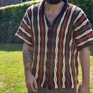 Puede incluir: Una camisa de manga corta con botones y dise&ntilde;o de ganchillo. La camisa presenta rayas verticales en marr&oacute;n, crema y naranja. El cuello y los pu&ntilde;os son de color marr&oacute;n oscuro. La camisa tiene cuello en V y est&aacute; abotonada.
