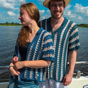 Op de afbeelding: Een gehaakt overhemd met korte mouwen en knoopsluiting, voorzien van verticale strepen in afwisselende kleuren blauwgroen en crème. Het kledingstuk heeft een kraag en een gestructureerd, opengewerkt breipatroon met een herhalend geometrisch ontwerp.