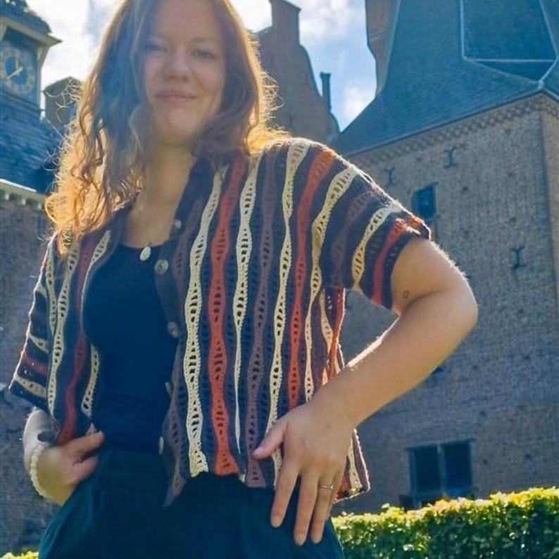 Puede incluir: C&aacute;rdigan de crochet de manga corta con botones, con rayas verticales en marr&oacute;n, crema, naranja y marr&oacute;n oscuro. El c&aacute;rdigan se lleva sobre un top negro y se combina con una falda verde oscuro. Con cierre de bot&oacute;n.