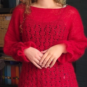 Op de afbeelding: Een rode, open gebreide trui met een boothals en lange mouwen. De mouwen hebben een gerimpelde manchet. De trui heeft een getextureerde, fuzzy uitstraling. Het onderste deel van de trui heeft een bloemenpatroon.