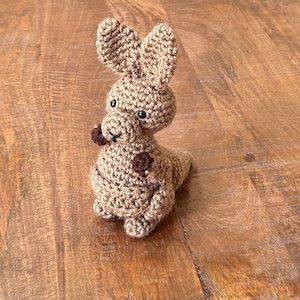 May include: A crocheted kangaroo toy with brown fur and black eyes. The kangaroo is sitting on a wooden surface.