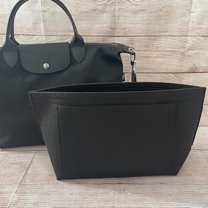 May include: A black handbag with a matching black felt organizer insert. The handbag has a flap closure and a top handle. The organizer has a rectangular shape with a pocket on the front. The background is a light brown wooden surface.