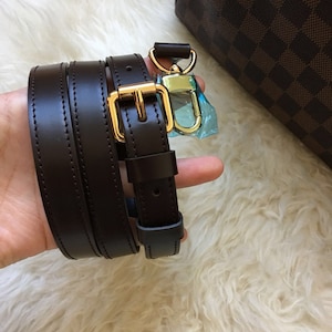 May include: A brown leather strap with a gold metal buckle and a gold metal hook. The strap is folded in half and is being held by a hand.