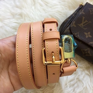 May include: A tan leather strap with gold hardware and stitching. The strap is folded in half and has a buckle and a hook.